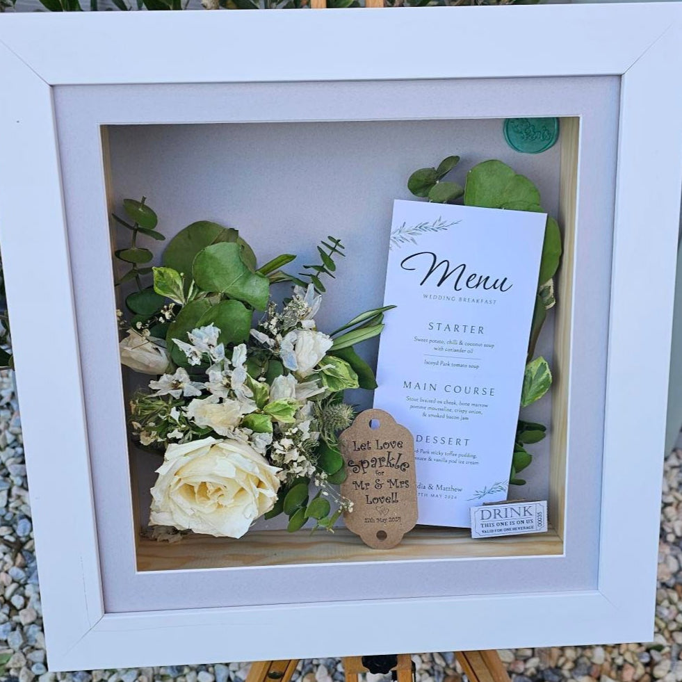 White framed shadow box with flowers and a card on a gravel surface