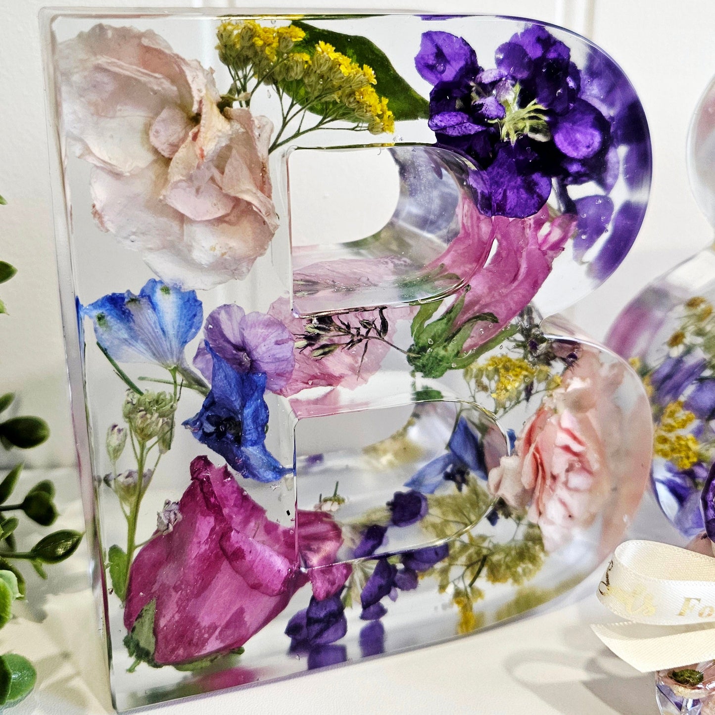 Decorative letter 'B' made of preserved flowers in resin on a white background
