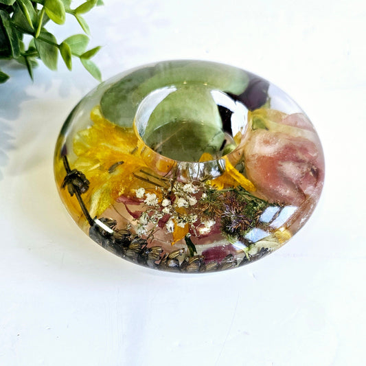 Round resin tealight holder i with embedded flowers and leaves on a white surface with greenery in the background